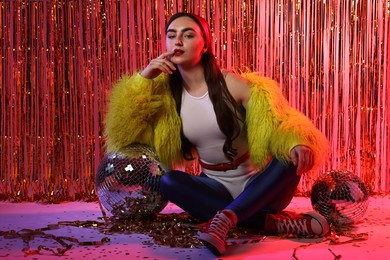 Photo of Beautiful young woman wearing retro outfit with tinsel and disco balls against golden foil curtain in neon lights