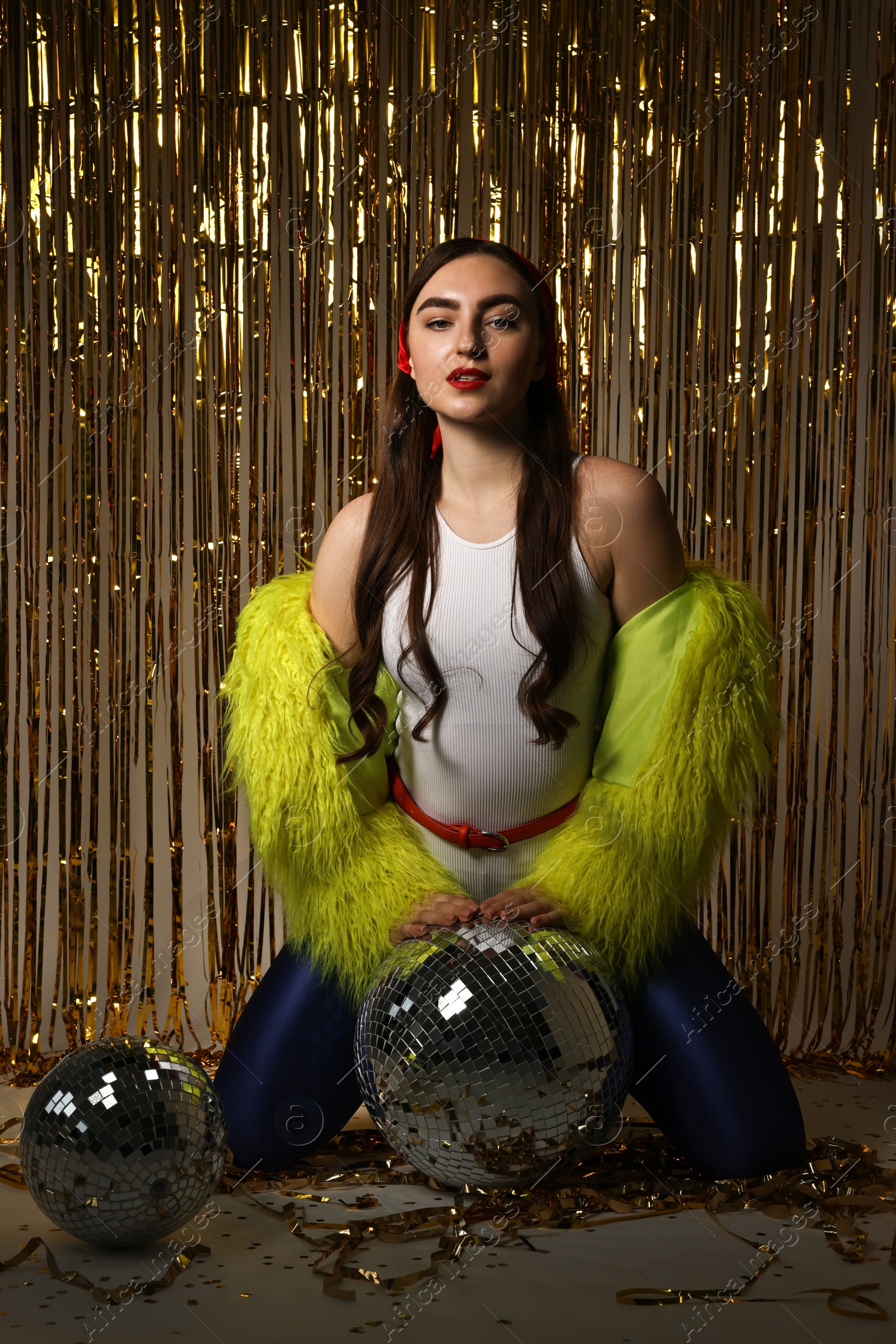 Photo of Beautiful young woman in retro outfit with tinsel and disco balls against golden foil curtain indoors