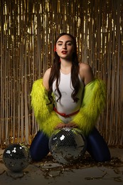 Photo of Beautiful young woman in retro outfit with tinsel and disco balls against golden foil curtain indoors