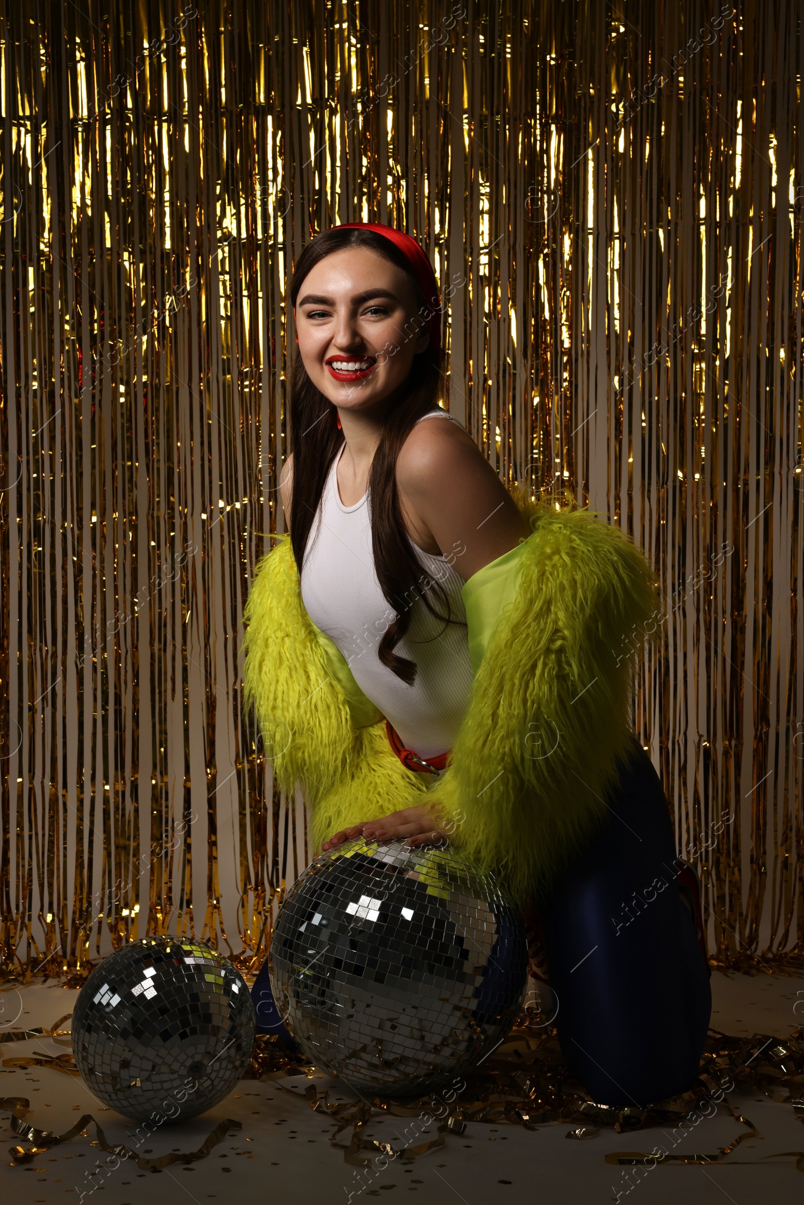 Photo of Beautiful young woman in retro outfit with tinsel and disco balls against golden foil curtain indoors