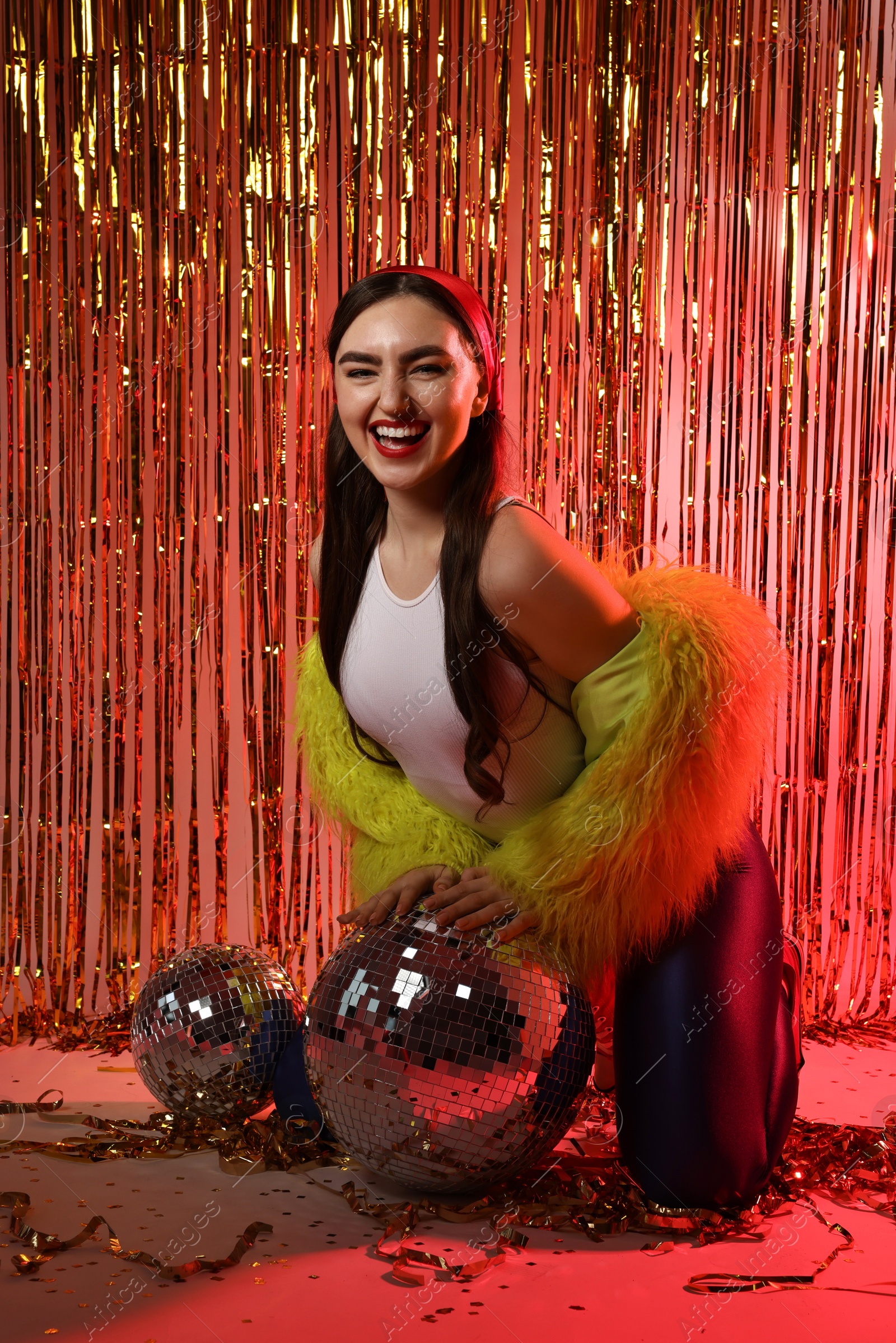 Photo of Beautiful young woman wearing retro outfit with tinsel and disco balls against golden foil curtain in neon lights