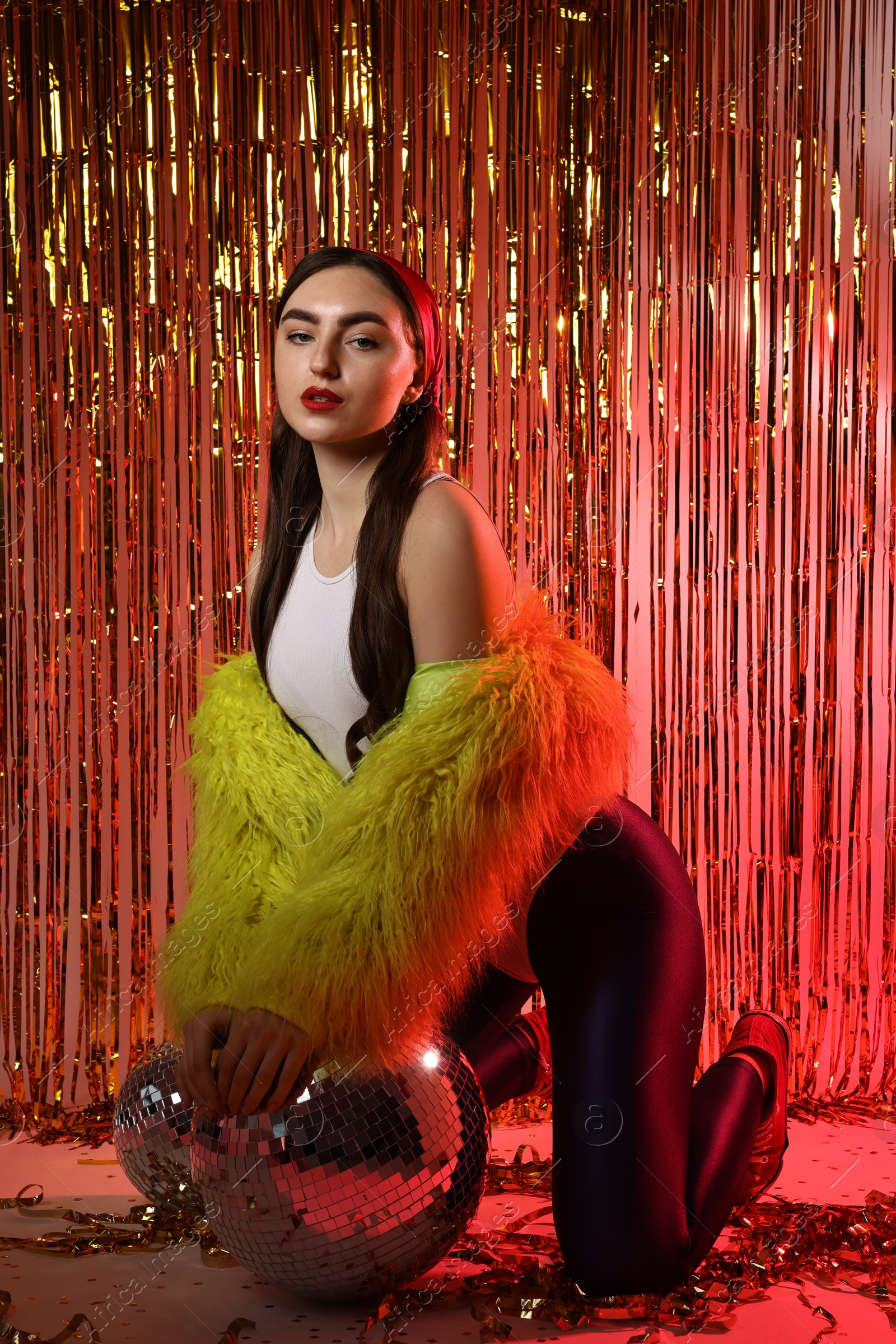 Photo of Beautiful young woman wearing retro outfit with tinsel and disco balls against golden foil curtain in neon lights