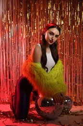 Photo of Beautiful young woman wearing retro outfit with tinsel and disco balls against golden foil curtain in neon lights