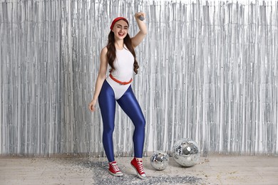 Photo of Beautiful young woman in retro outfit with microphone near silver foil curtain indoors