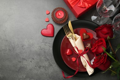 Romantic place setting for Valentine's day. Tableware, cutlery, candies, roses and decorative hearts on grey textured table, flat lay. Space for text