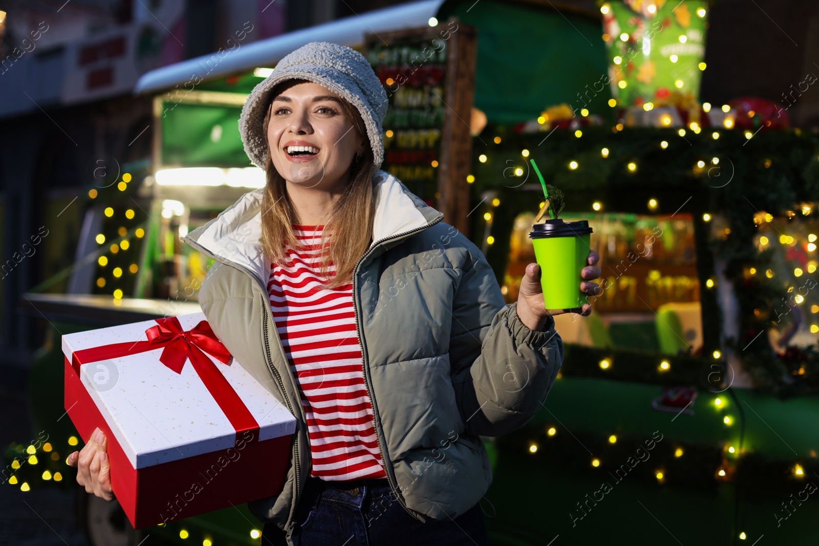 Photo of Happy woman with Christmas gift and paper cup near festive bus outdoors