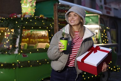 Photo of Happy woman with Christmas gift and paper cup near festive bus outdoors. Space for text