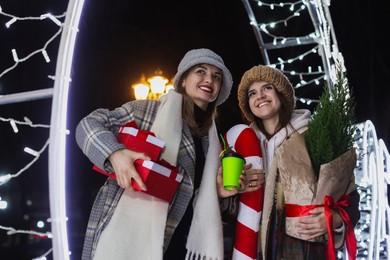 Photo of Happy friends with gifts and thuja tree near Christmas decoration at night outdoors