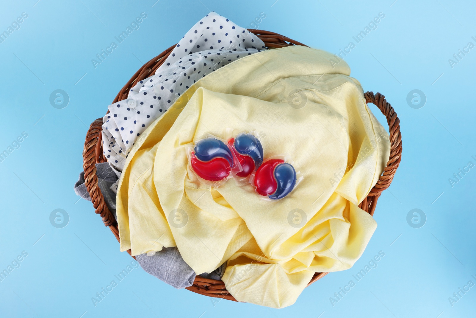 Photo of Detergent capsules and laundry in basket on light blue background, top view