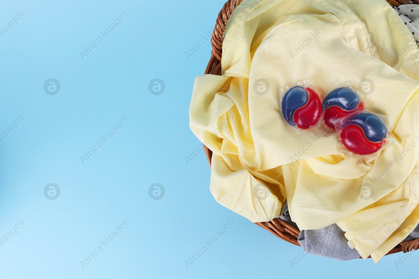 Photo of Detergent capsules and laundry in basket on light blue background, top view. Space for text