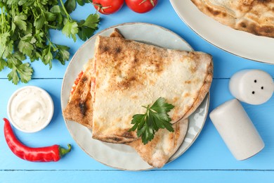 Photo of Halves of tasty calzone, tomato, chili pepper, parsley and sauce on light blue table, flat lay