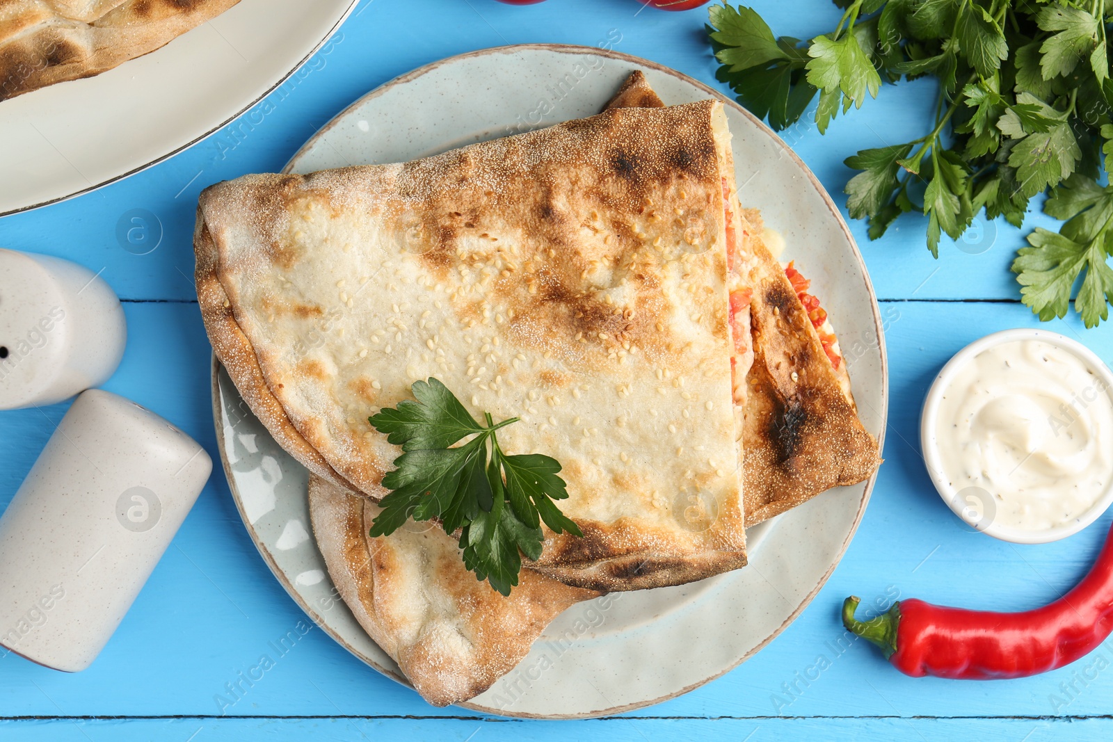 Photo of Halves of tasty calzone, tomato, chili pepper, parsley and sauce on light blue table, flat lay