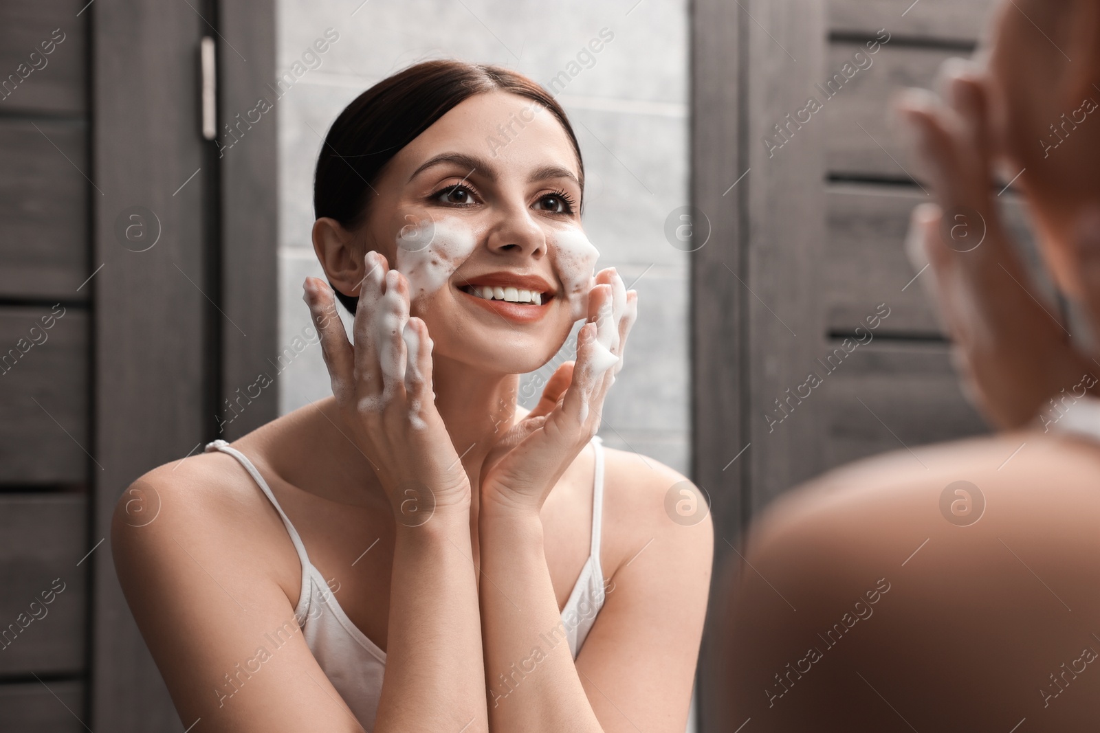 Photo of Woman washing her face with cleansing foam near mirror in bathroom