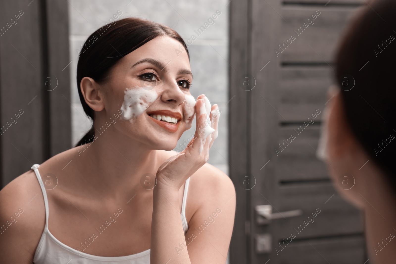 Photo of Woman washing her face with cleansing foam near mirror in bathroom