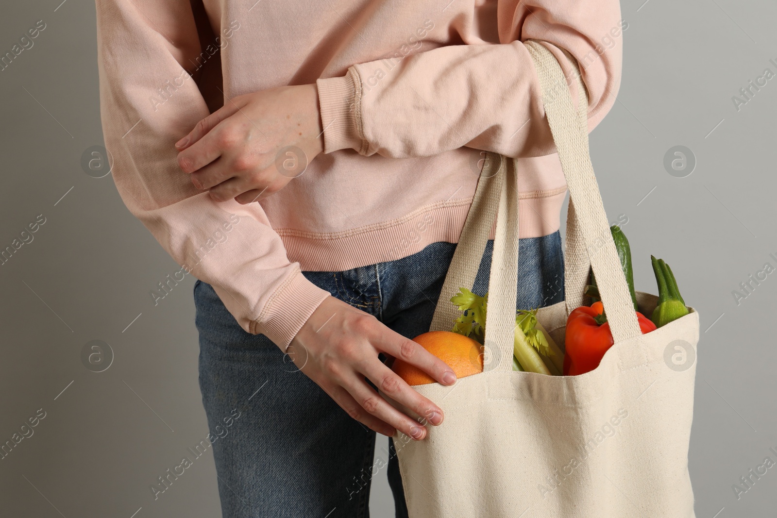 Photo of Woman holding shopper bag with groceries on light grey background, closeup. Eco friendly lifestyle