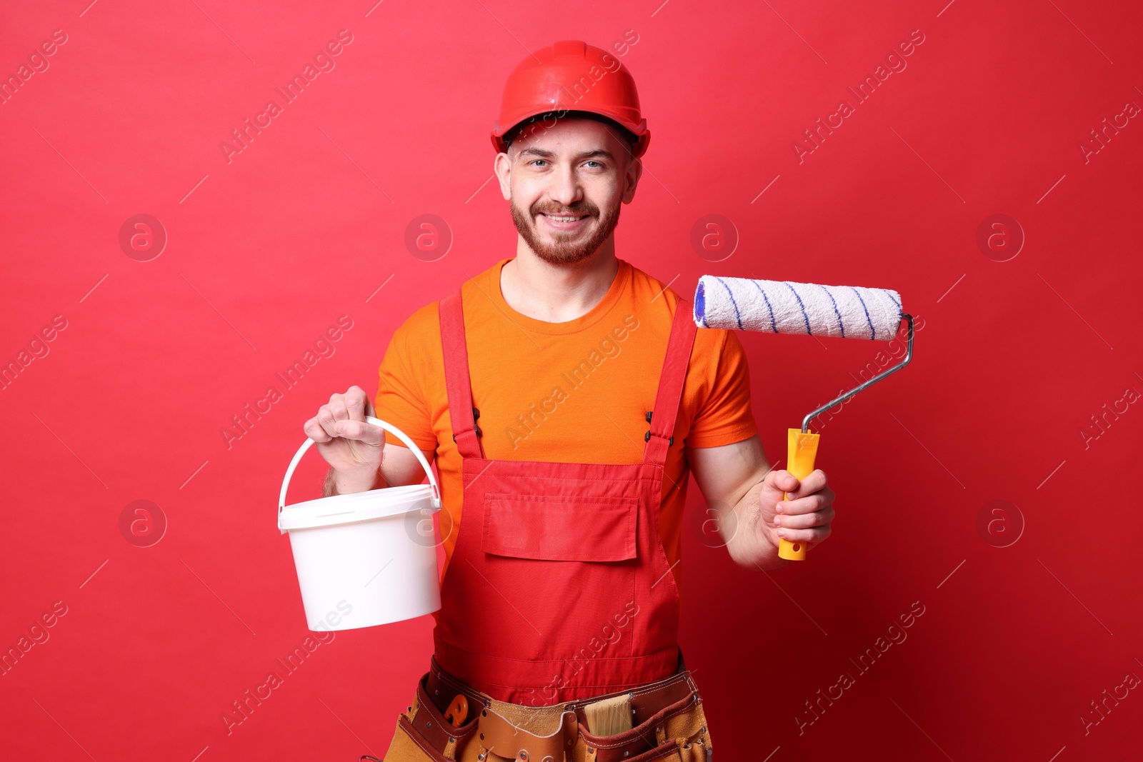 Photo of Professional painter with roller and bucket of paint on red background