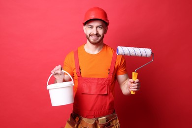 Photo of Professional painter with roller and bucket of paint on red background