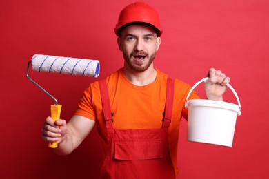 Photo of Emotional painter with roller and bucket of paint on red background