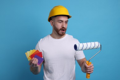 Photo of Emotional man wearing hardhat with roller and color samples on blue background