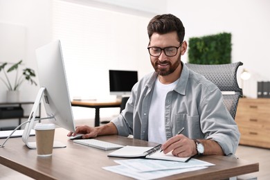 Photo of Man taking notes while working on computer at table in office