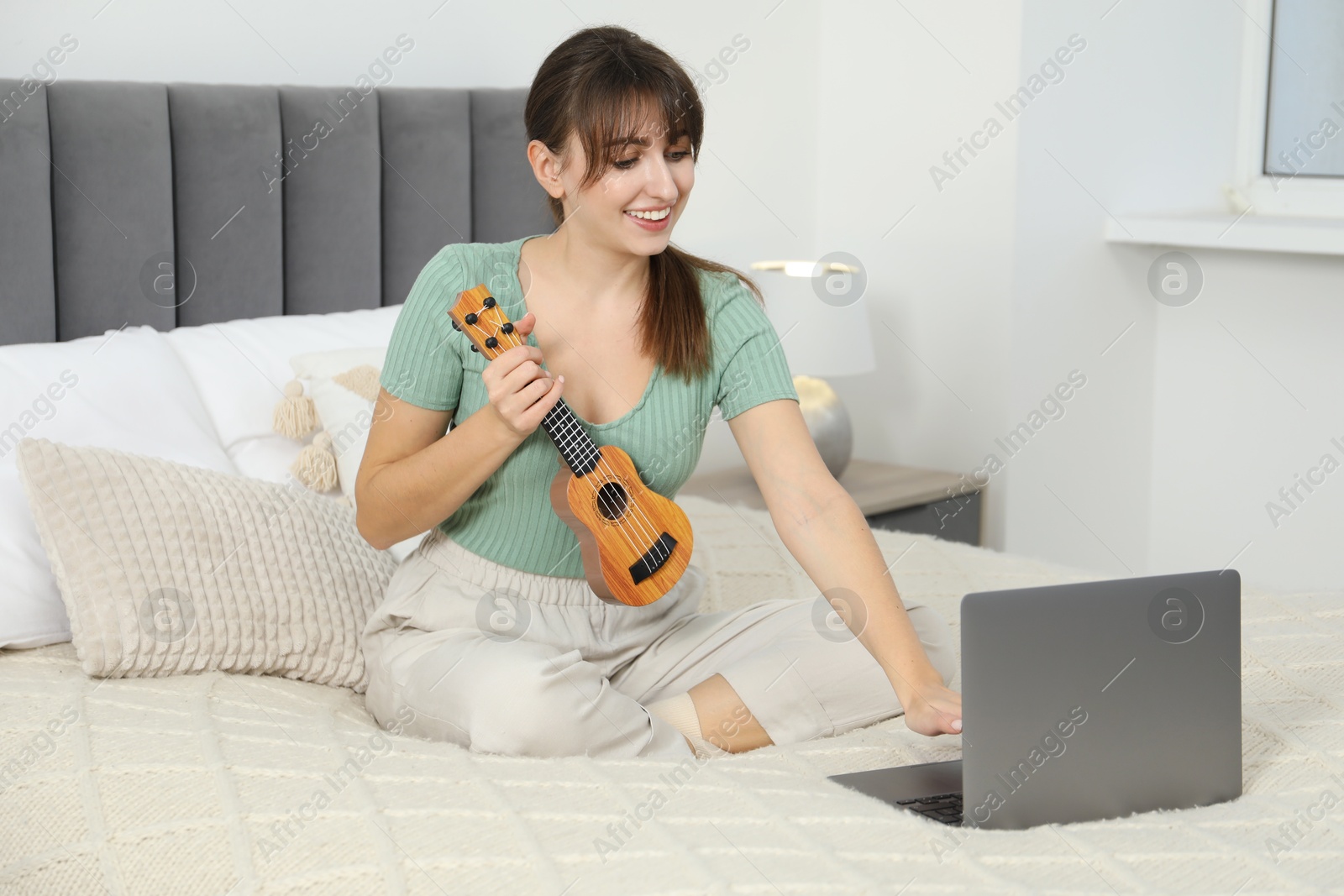 Photo of Happy woman learning to play ukulele with online music course on bed at home