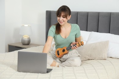 Photo of Happy woman learning to play ukulele with online music course on bed at home