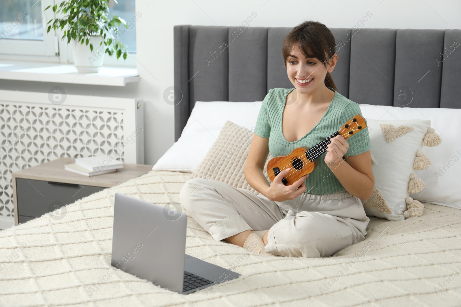 Photo of Happy woman learning to play ukulele with online music course on bed at home