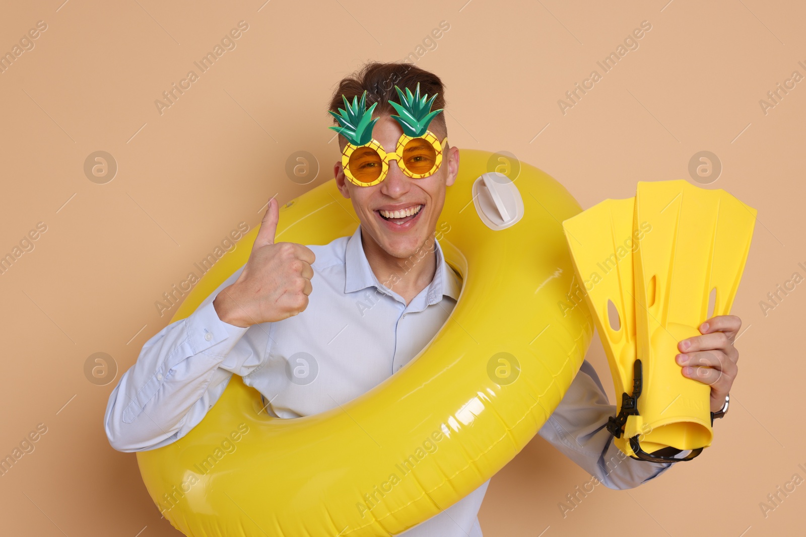 Photo of Businessman with inflatable ring, funny sunglasses and flippers showing thumbs up on beige background