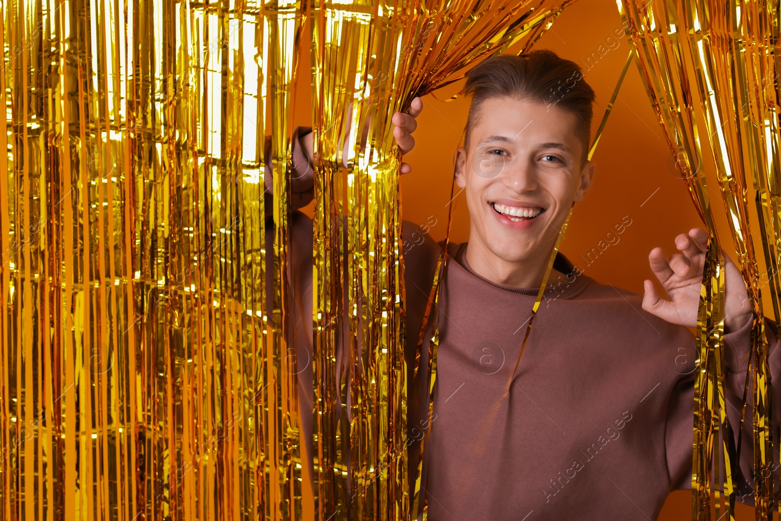 Photo of Happy young man looking out through golden foil curtain against orange background