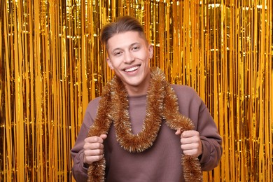 Photo of Happy young man with bright tinsel against golden foil curtain