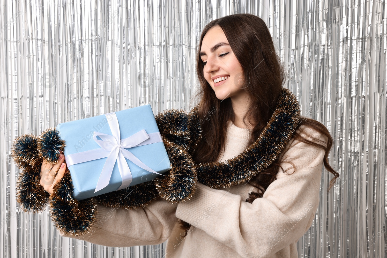 Photo of Happy young woman with tinsel and gift box against silver foil curtain