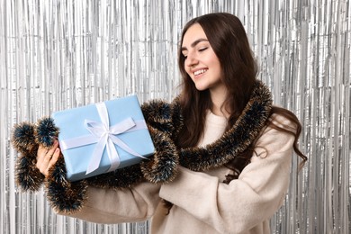 Photo of Happy young woman with tinsel and gift box against silver foil curtain