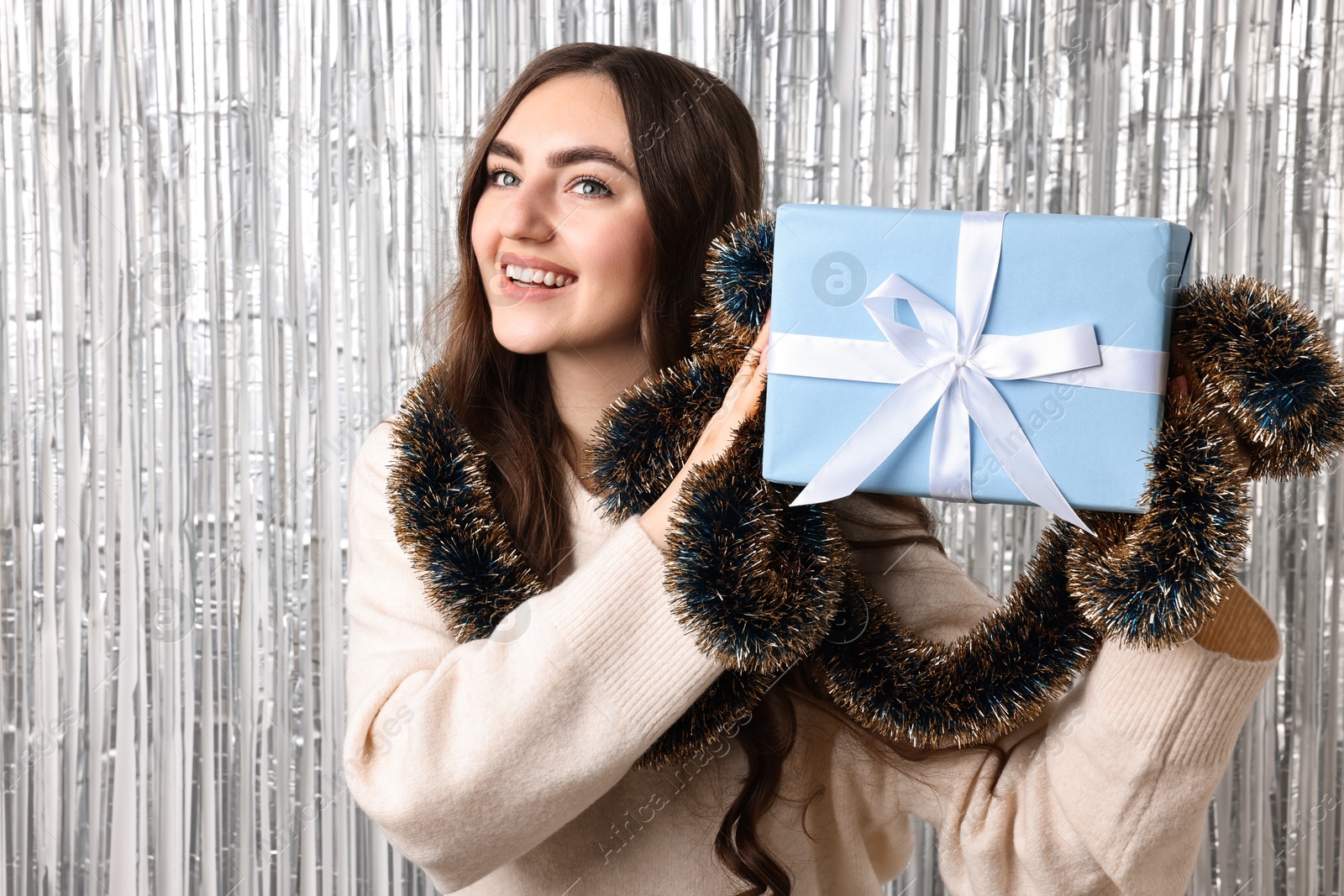 Photo of Happy young woman with tinsel and gift box against silver foil curtain