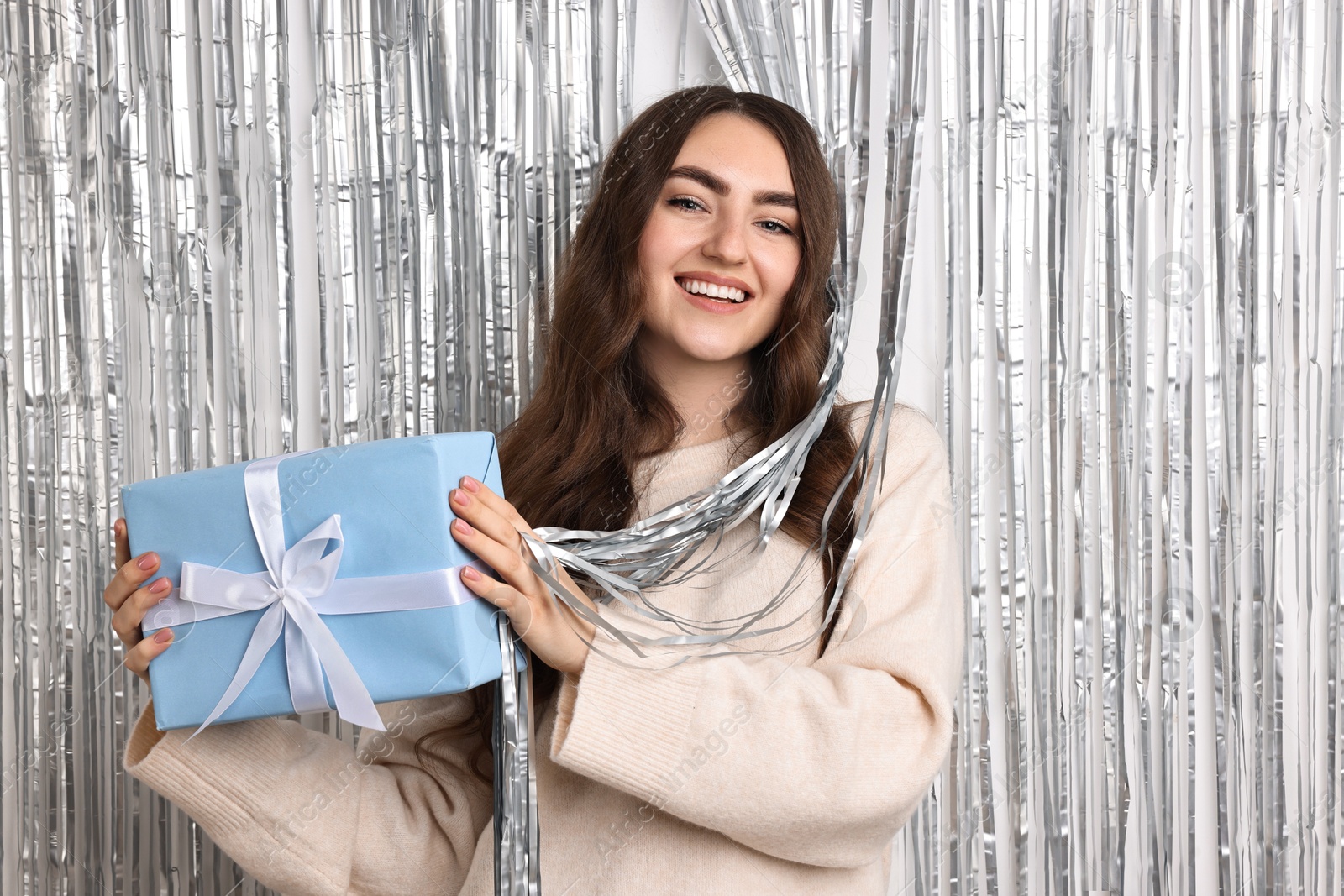 Photo of Happy young woman with gift box near silver foil curtain