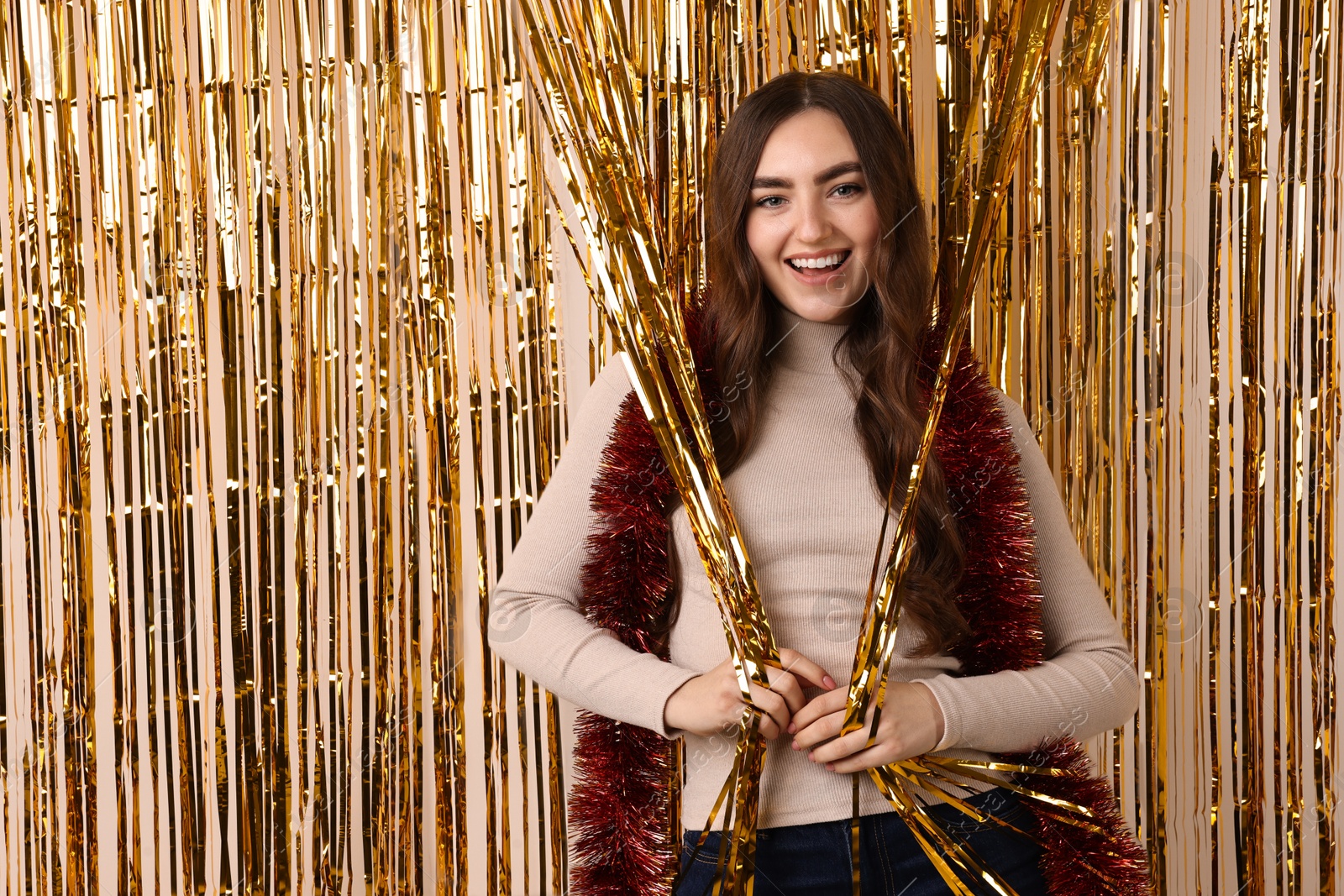 Photo of Happy young woman with tinsel near golden foil curtain against beige background