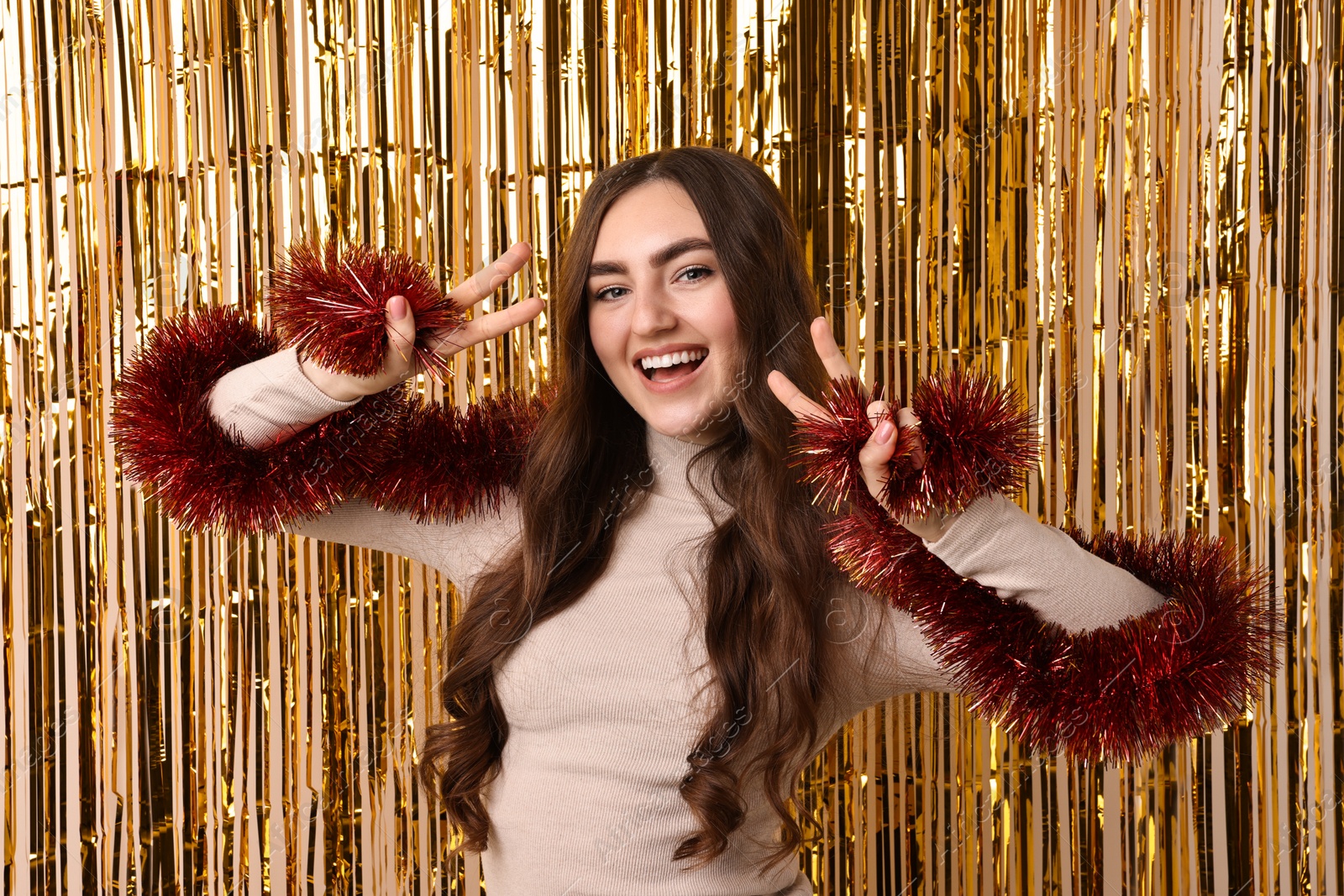 Photo of Happy young woman with tinsel showing V-signs against golden foil curtain