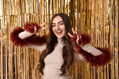 Photo of Happy young woman with tinsel showing V-signs against golden foil curtain