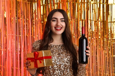 Photo of Happy young woman with gift box and bottle of wine against foil curtain