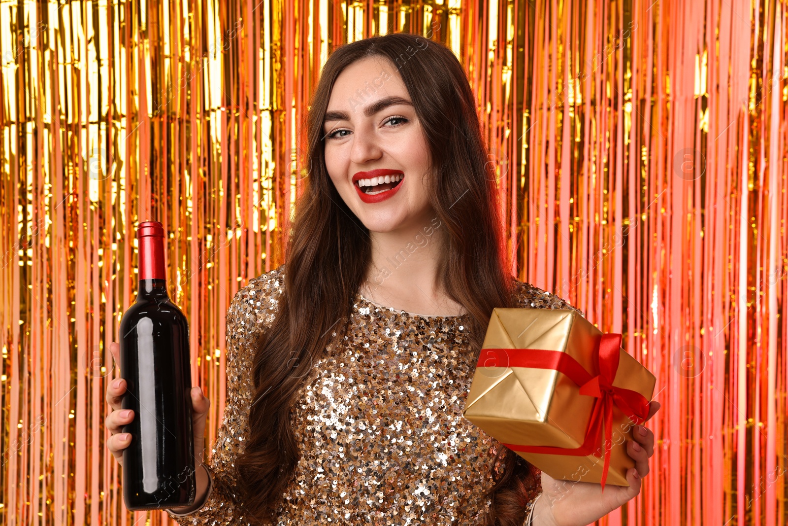 Photo of Happy young woman with gift box and bottle of wine against foil curtain