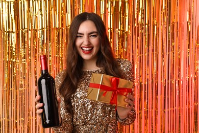 Photo of Happy young woman with gift box and bottle of wine against foil curtain