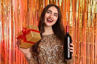 Photo of Happy young woman with gift box and bottle of wine against foil curtain