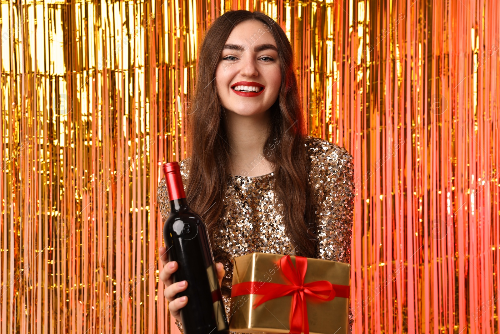 Photo of Happy young woman with gift box and bottle of wine against foil curtain