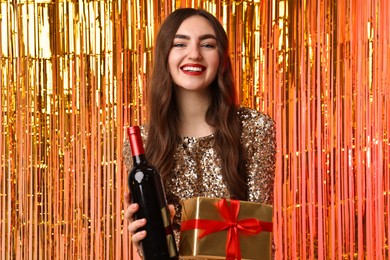 Photo of Happy young woman with gift box and bottle of wine against foil curtain