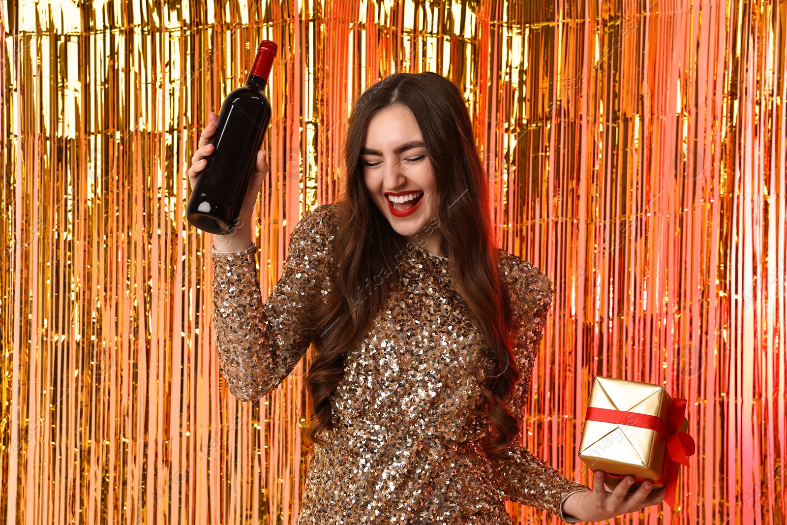 Photo of Happy young woman with gift box and bottle of wine against foil curtain