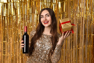 Photo of Happy young woman with gift box and bottle of wine against golden foil curtain