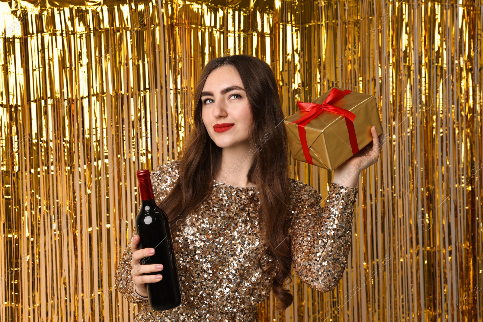 Photo of Young woman with gift box and bottle of wine against golden foil curtain