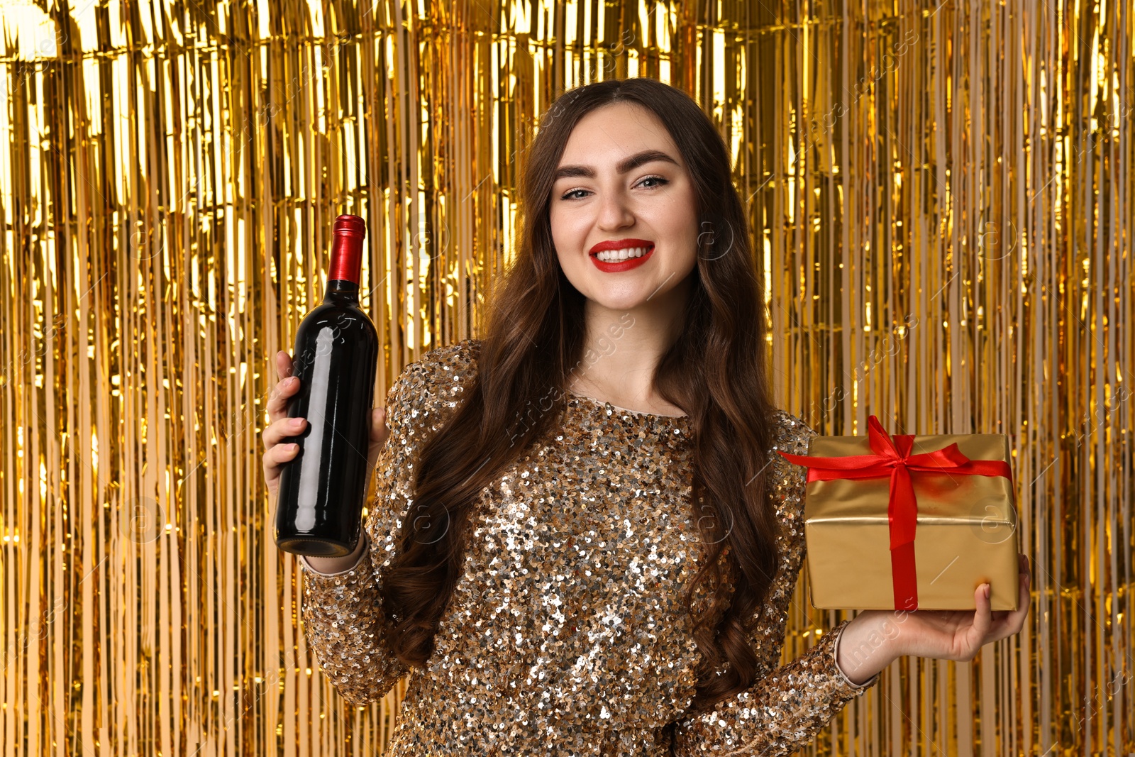 Photo of Happy young woman with gift box and bottle of wine against golden foil curtain