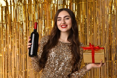 Photo of Happy young woman with gift box and bottle of wine against golden foil curtain