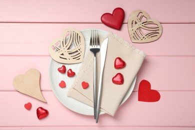 Photo of Romantic place setting for Valentine's day. Cutlery, napkin, plate and decorative hearts on pink wooden table, flat lay