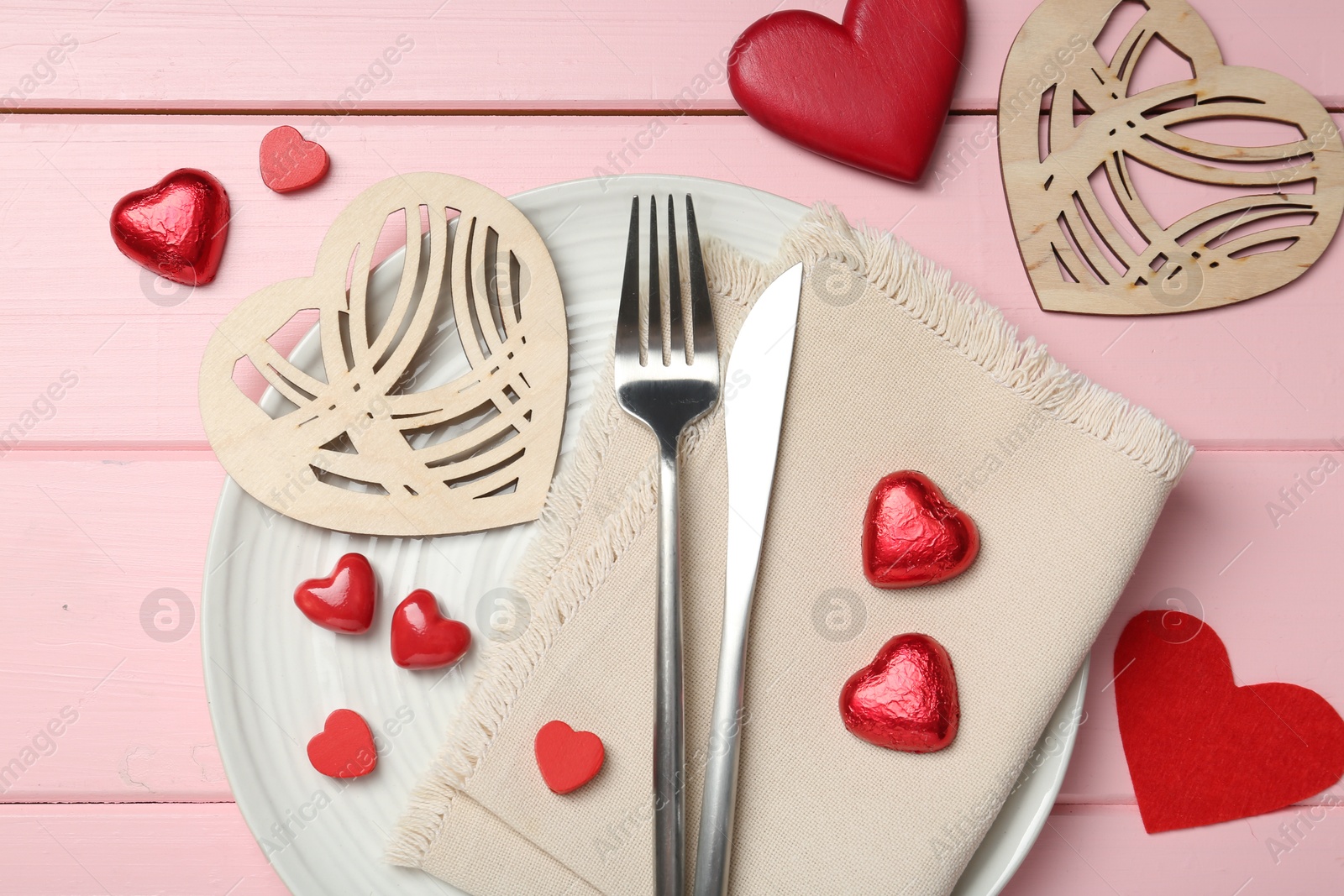 Photo of Romantic place setting for Valentine's day. Cutlery, napkin, plate and decorative hearts on pink wooden table, flat lay